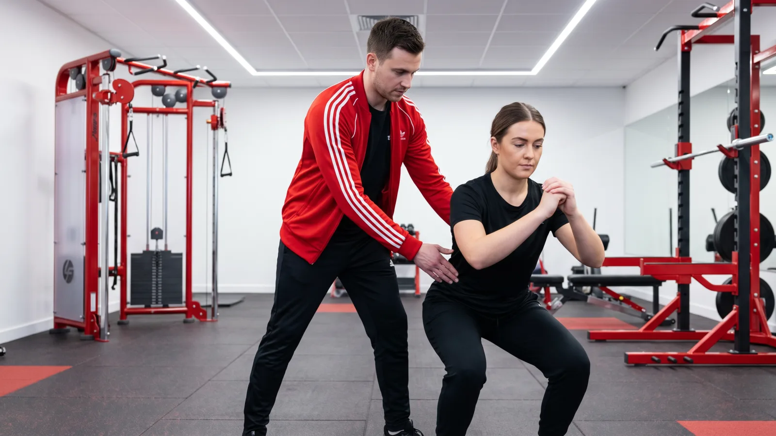 Ein Trainer und eine Klientin führen eine Kniebeuge durch, passend für Physiotherapie und Training kombiniert.